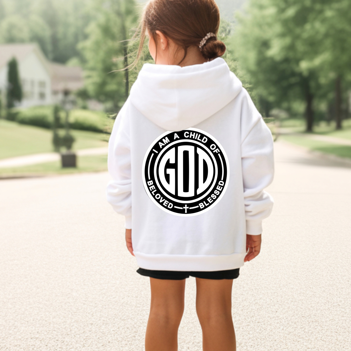 A child viewed from behind wearing a white pullover hoodie with a large black circular graphic on the back that reads 'I AM A CHILD OF GOD' and 'BLESSED / BLESSED.' The child is standing outdoors on a paved path with trees and a house blurred in the background.