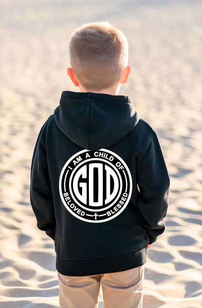 A child viewed from behind wearing a black hoodie with a large white circular graphic on the back that reads 'I AM A CHILD OF GOD' and 'BELoved + BLESSED'. The child is standing outdoors on sandy ground.
