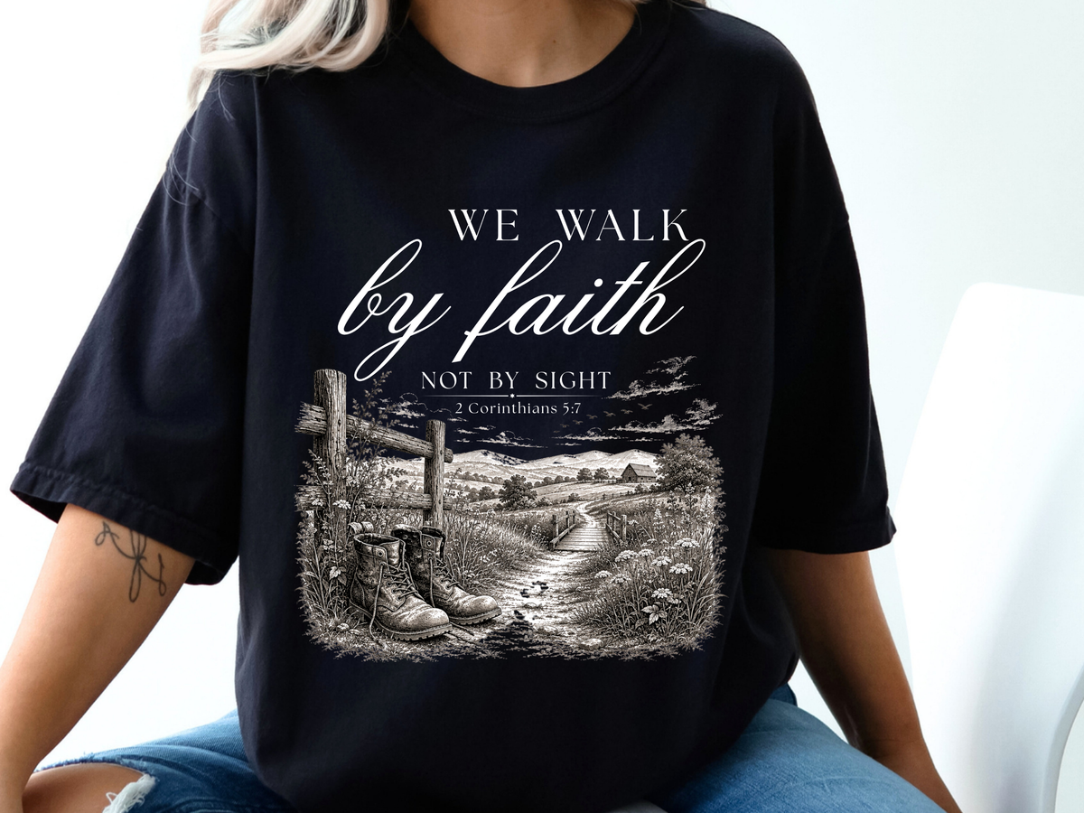 A Christian t-shirt with an image of a path cutting through fields and trees with a barn in the background and a fence with a pair of boots sitting in front, the words We walk by faith not by sight 2 Corinthians 5:7 are just above the image.