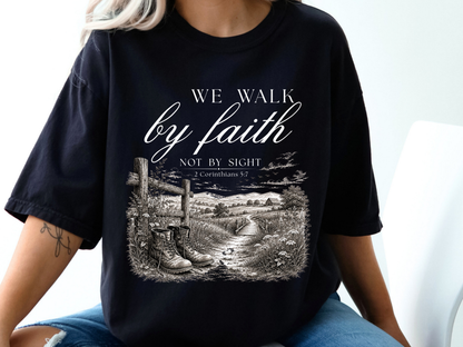 A Christian t-shirt with an image of a path cutting through fields and trees with a barn in the background and a fence with a pair of boots sitting in front, the words We walk by faith not by sight 2 Corinthians 5:7 are just above the image.