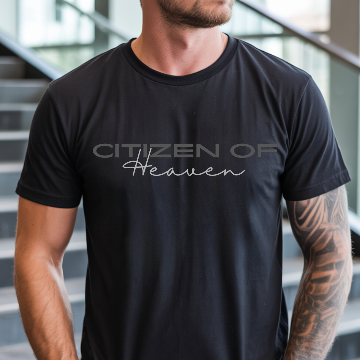 A man wearing a black crew-neck t-shirt with the text 'CITIZEN OF Heaven' printed across the chest in gray and white, standing in front of a blurred staircase background.