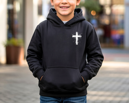 A smiling young boy wearing a black pullover hoodie with a white distressed cross graphic on the left chest, standing outdoors on a paved street with a blurred background.