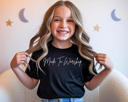A smiling young girl with long blonde hair wearing a black t-shirt with 'Made To Worship' printed in white script, posing indoors against a light wall with moon and star decorations.