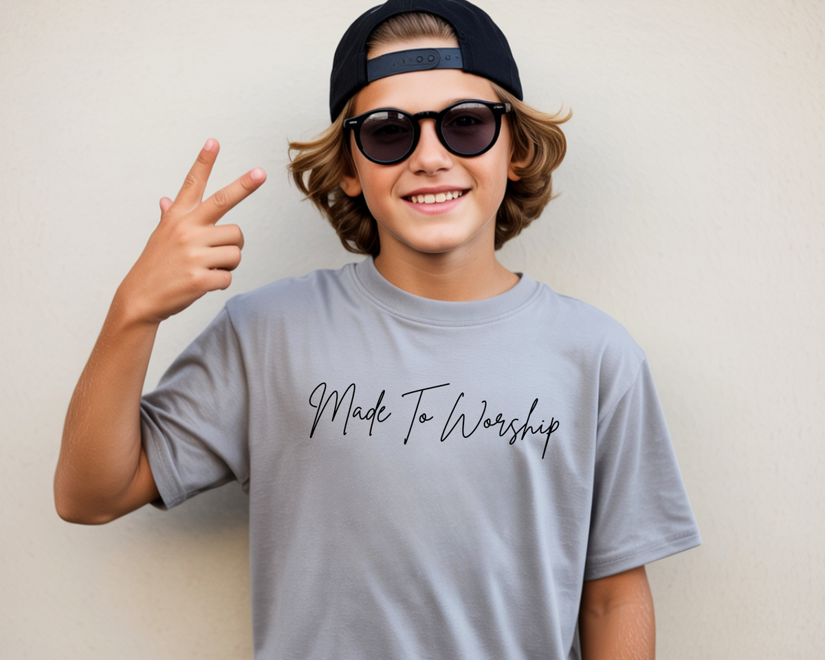 A smiling young man wearing a black baseball cap backwards, black sunglasses, and a light gray t-shirt with 'Made To Worship' printed in black script across the chest. He is posing with a peace sign against a plain beige wall.
