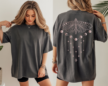 A woman modeling an oversized dark gray washed T-shirt shown from both front and back. The front is plain, while the back features a large pale pink moon-phase dragonfly graphic.