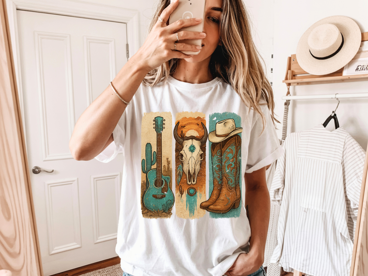 A woman takes a mirror selfie wearing a white graphic t-shirt with a western-themed print featuring a turquoise guitar, a brown skull, and a cowboy boot. The background shows a room with a wooden door, a hat on a shelf, and a striped shirt hanging on a rack.