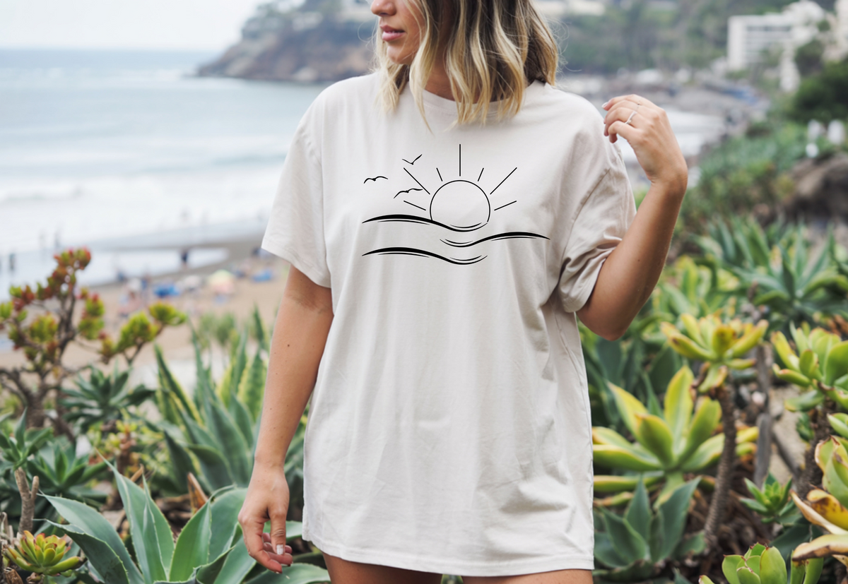 A woman standing on a beach wearing an oversized ivory t-shirt the image on the front is a simple line drawing of waves and a sun with birds flying around the sun.