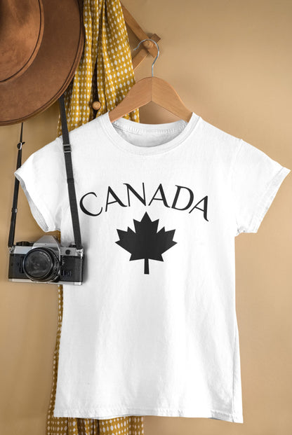A white short-sleeve T-shirt hanging on a wooden hanger against a warm beige wall, printed with the word CANADA in black serif letters above a black maple leaf graphic. A brown hat, yellow-and-white checkered fabric, and a vintage camera are styled around it as props.